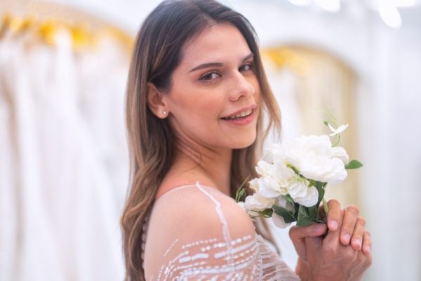 Beautiful young brunette woman choosing wedding dress in a bridal salon smelling flower