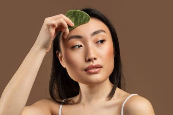 Sensual asian lady doing daily skin care routine, massaging facial skin with jade gua sha scrapper