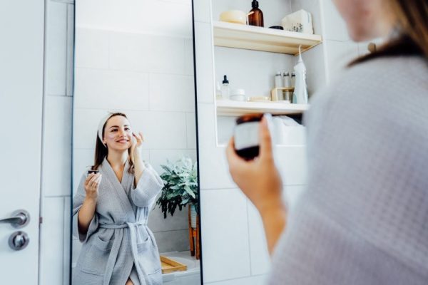 Young woman in bathrobe looking in mirror and applying facial natural cosmetic clay mask on face